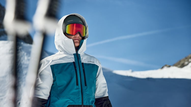 Person mit blauer und weißer Winterjacke und hochgezogener Kapuze sowie Skibrille vor einer verschneiten Berglandschaft unter klarem, blauem Himmel