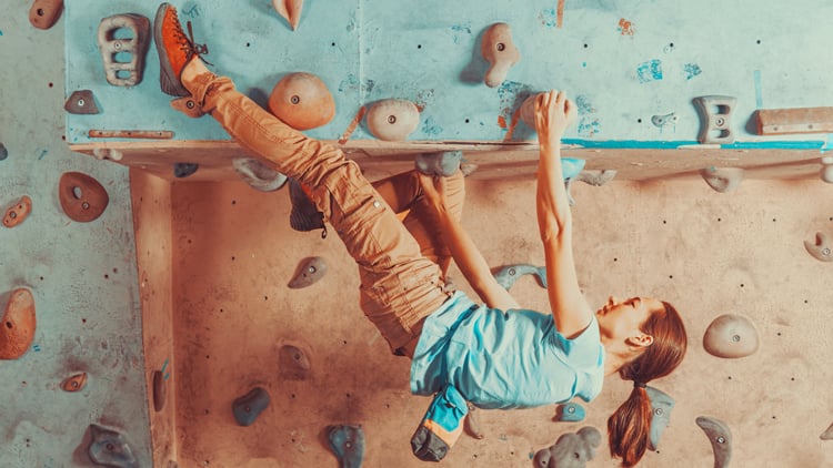 Eine Person beim Indoor-Klettern an einer überhängenden Wand. Sie trägt ein blaues Shirt, beige Hose und Kletterschuhe und nutzt farbige Griffe in verschiedenen Formen und Größen mit Händen und Füßen.