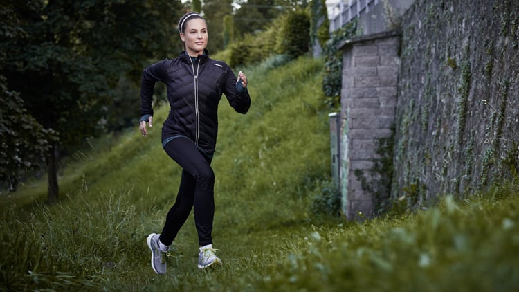Person beim Joggen auf einem grasbewachsenen Weg, gekleidet in schwarze Jacke und Leggings sowie weiß-graue Laufschuhe mit grünen Akzenten. Im Hintergrund sind Bäume und eine mit Vegetation bewachsene Steinmauer zu sehen