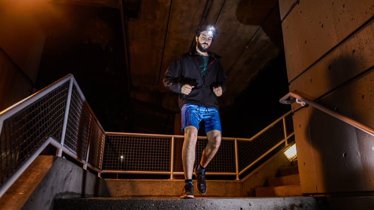 Laufberater Eine Person läuft nachts eine Treppe in städtischer Umgebung hinunter. Sie trägt eine Stirnlampe, eine dunkle Jacke und blaue Shorts. Betonwände und Metallgeländer umgeben die Szene, die von künstlichem Licht beleuchtet wird