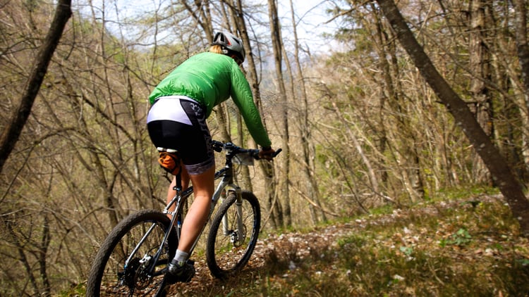 Person auf einem Mountainbike fährt auf einem Waldweg; trägt grüne Jacke, schwarz-weiße Shorts und Helm.