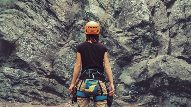 Eine Person mit einem orangefarbenen Helm und Kletterausrüstung steht vor einer felsigen Klippe