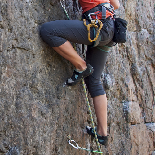 Eine Person beim Felsklettern an einer senkrechten Wand. Sie trägt Kletterausrüstung mit Gurt, Kletterschuhen sowie mehreren Karabinern und Seilen. Die Beine sind angewinkelt, während sie die Wand hinaufsteigt.