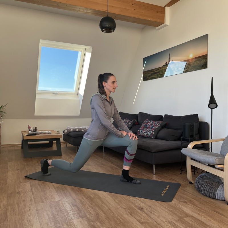 Person macht einen Ausfallschritt auf einer Yogamatte im Wohnzimmer mit Holzfußboden