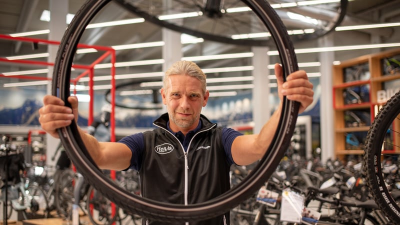 Ein Hervis Mitarbeiter hält ein Fahrradlaufrad hoch. Im Hintergrund sind mehrere Fahrräder sowie Fahrradzubehör auf Regalen und Ständern zu sehen.