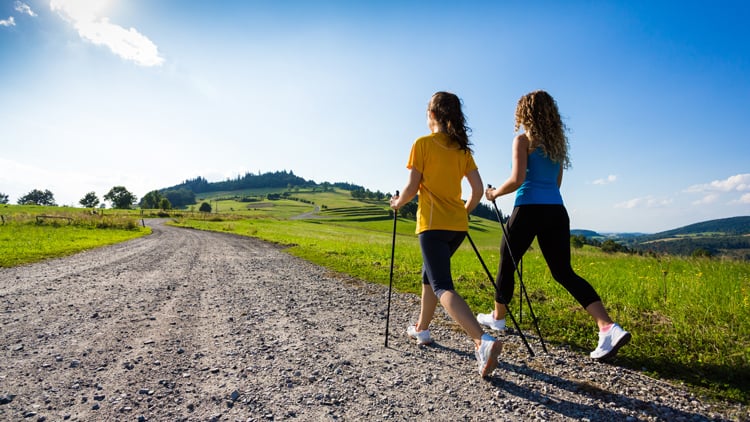 Zwei Personen beim Nordic Walking auf einem Kiesweg in hügeliger Landschaft. Eine trägt ein gelbes Shirt, die andere ein blaues Top, beide schwarze Caprihosen. Umgeben von grüner Natur und leicht bewölktem Himmel
