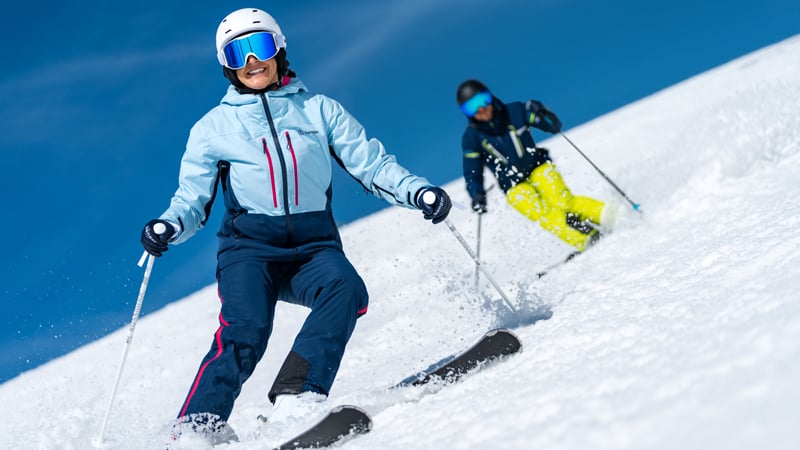 Zwei Skifahrer fahren bei Sonnenschein eine verschneite Piste hinunter.