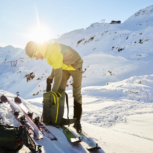 Person in Skiausrüstung steht auf einem verschneiten Berghang, beugt sich über einen Rucksack am Boden. Zwei Paar Skier und Skistöcke liegen im Schnee daneben. Die Sonne scheint hell und beleuchtet die winterliche Berglandschaft im Hintergrund