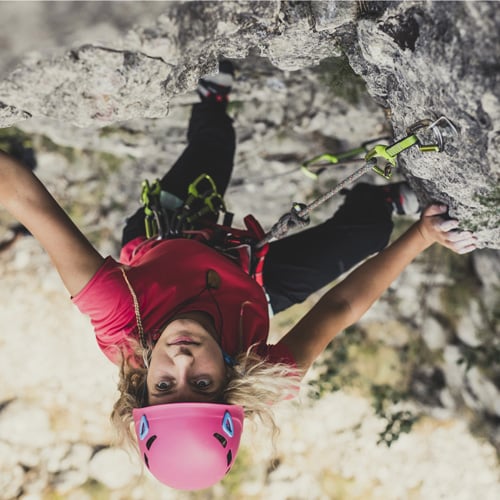 Person beim Klettern an einer Felswand, gesichert mit Kletterausrüstung. Sie trägt einen pinken Helm, ein pinkes Shirt und schwarze Hose. Im Hintergrund sind Felsen und etwas Vegetation zu sehen