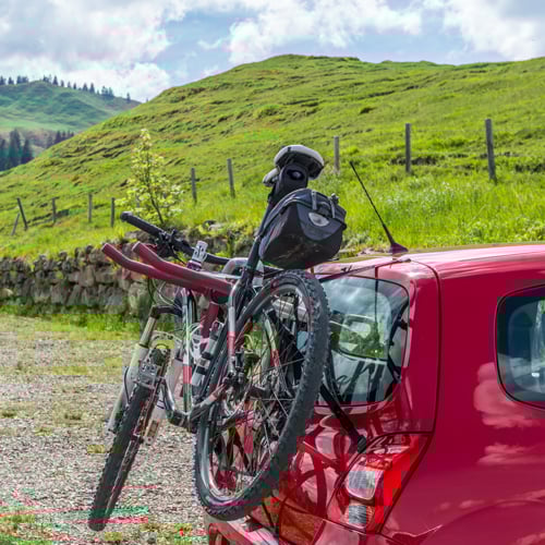 Rotes Auto mit Heckklappenträger, auf dem ein Fahrrad montiert ist; im Hintergrund grüne Hügel, eine Steinmauer und teils bewölkter Himmel in ländlicher Umgebung.