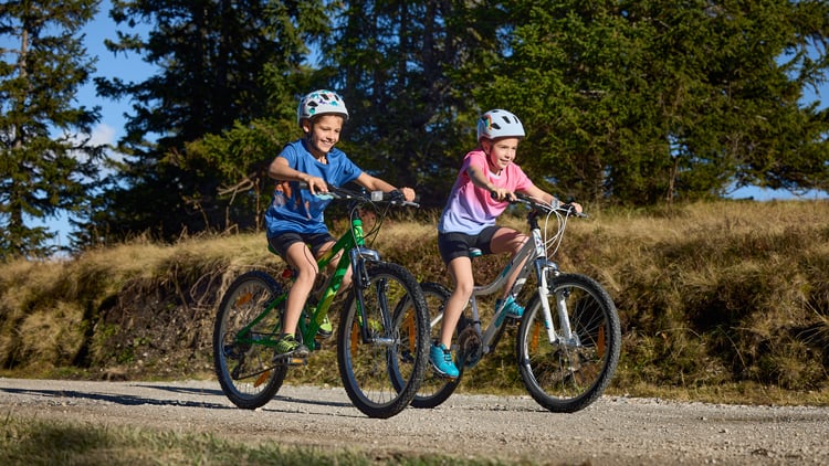 Zwei Kinder fahren mit Fahrrädern auf einem unbefestigten Weg durch eine grüne Landschaft mit Bäumen und Gras im Hintergrund; links ein grünes Fahrrad, rechts ein weißes, beide Fahrer tragen Helme und Freizeitkleidung.