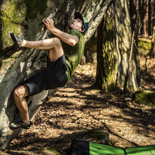 Eine Person beim Bouldern an einer Felswand im Freien. Sie trägt ein grünes Shirt, schwarze Shorts und Kletterschuhe. Unter ihr liegt ein Crashpad zur Absicherung. Im Hintergrund sind Bäume und Sonnenlicht, das durch den Wald fällt.