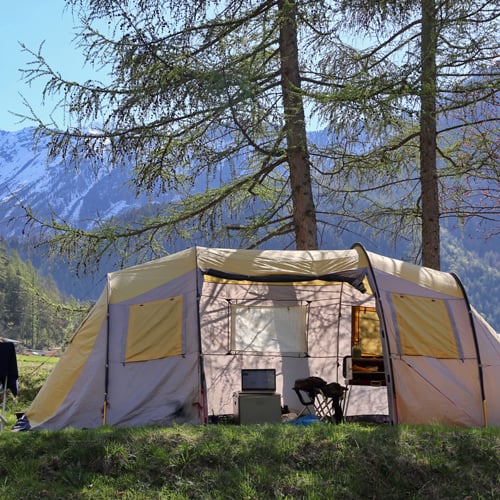 Großes beige-gelbes Campingzelt mit offenem Eingang, in dem Campingausrüstung wie Stühle und ein kleiner Tisch sichtbar sind. Das Zelt steht auf einer Wiese, umgeben von hohen Bäumen, mit schneebedeckten Bergen im Hintergrund.