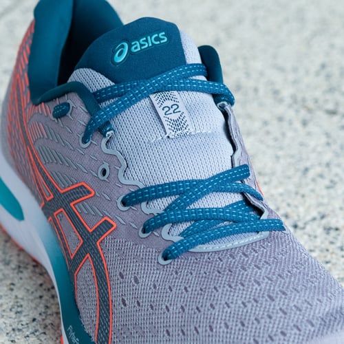 Nahaufnahme eines grauen Asics-Laufschuhs mit blauen Schnürsenkeln, blauen und orangefarbenen Akzenten. Das Asics-Logo ist auf der Zunge und der Seite sichtbar. Auf der Zunge befindet sich zudem die Zahl ‚22‘, die auf das Modell hinweist
