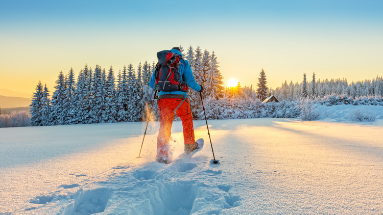 Eine Person beim Schneeschuhwandern in einer verschneiten Landschaft bei Sonnenuntergang. Sie trägt eine blaue Jacke, rote Hose, einen Rucksack und benutzt Skistöcke. Im Hintergrund sind schneebedeckte Bäume und ein klarer Himmel mit untergehender Sonne zu sehen