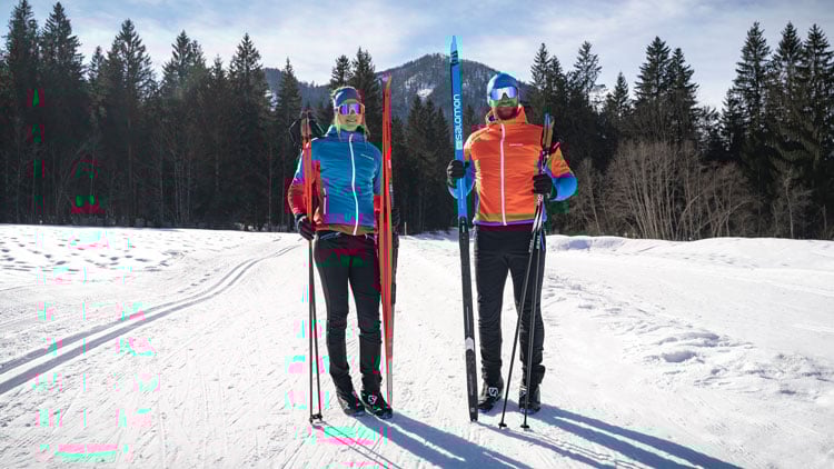 Zwei Personen stehen nebeneinander in einer verschneiten Landschaft, beide mit Skistöcken und Skibekleidung. Die linke Person trägt eine blaue Jacke mit roten Akzenten, schwarze Hose und reflektierende Sonnenbrille. Die rechte Person trägt eine orangefarbene Jacke mit blauen Akzenten, ebenfalls schwarze Hose und reflektierende Sonnenbrille. Im Hintergrund sind verschneite Böden und hohe Nadelbäume zu sehen