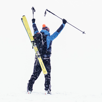 Person steht im Schnee mit dem Rücken zur Kamera, trägt eine blaue Winterjacke und eine orangefarbene Mütze. Gelbe Skier sind am Rücken befestigt, Skistöcke werden mit erhobenen Armen gehalten
