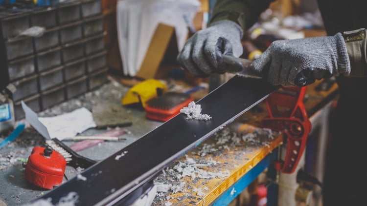 Person mit schwarzen Handschuhen bearbeitet ein Ski mit einem Werkzeug, um die Kante zu schärfen. Das Sportgerät ist mit roten Klemmen auf einer gelben Werkbank fixiert