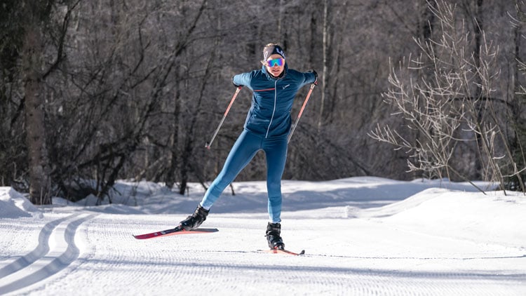 Eine Person beim Langlaufen auf einem verschneiten Weg, umgeben von Bäumen. Sie trägt blaue Skibekleidung mit Jacke, Hose, Handschuhen und Sonnenbrille. Mit Skistöcken in der Hand befindet sie sich in Bewegung, ein Ski ist angehoben