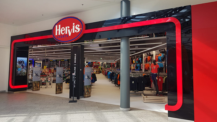 Der Eingang des Hervis Sports Stores im einem Einkaufszentrum Haid. Über dem Eingang befindet sich ein auffälliges rotes und weißes Logo mit der Aufschrift 'Hervis Sports'. Der Eingang ist von einem schwarz-roten Design umrahmt, mit Glastüren, die in den Store führen. Im Inneren sind verschiedene Sportbekleidung und -ausrüstung auf Regalen und Ständern ausgestellt.