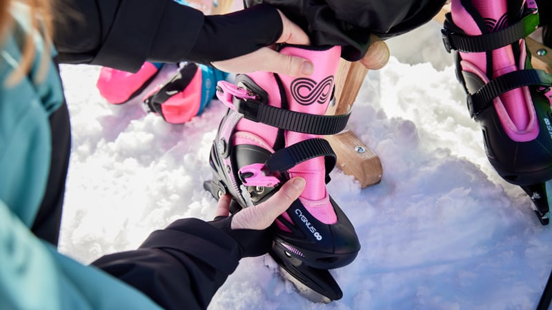 Ein Kind zieht pinke Eislaufschuhe an und bekommt dabei Hilfe beim Schließen der Schnallen