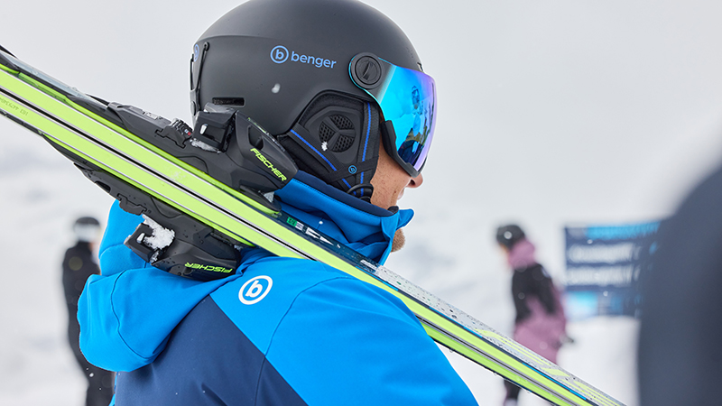 Ein Skifahrer in blauer Jacke trägt seine Ski über der Schulter, während er auf der Piste steht.