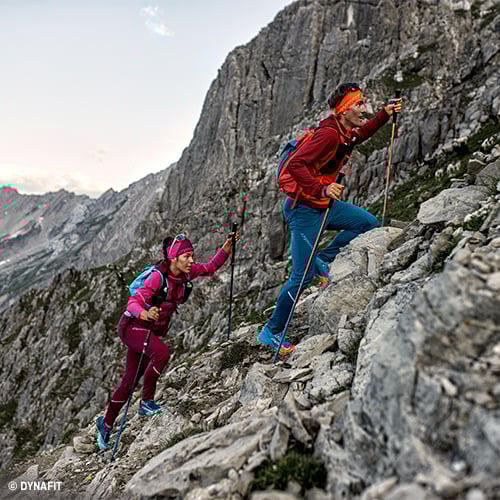 Zwei Personen wandern mit Trekkingstöcken und Outdoor-Ausrüstung einen felsigen Bergpfad hinauf; sie tragen wetterfeste Kleidung und Rucksäcke, im Hintergrund steile Felswände und teils bewölkter Himmel