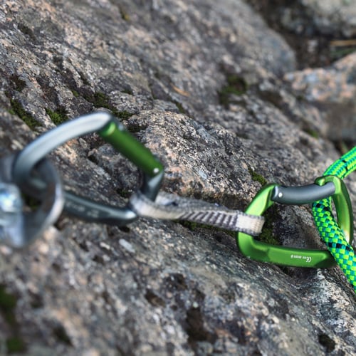 Nahaufnahme von Kletterausrüstung an einer Felswand: Zwei Karabiner, einer grün und einer silbern, sind mit einem Bandschlingensystem verbunden. Ein grünes Kletterseil ist am grünen Karabiner befestigt.