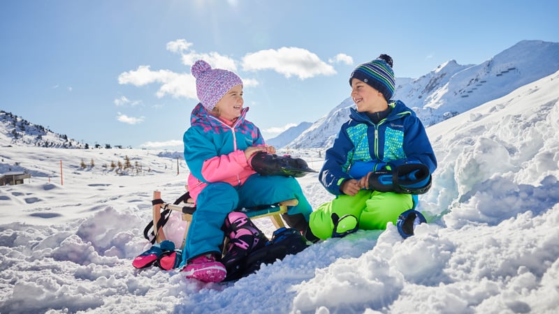 Zwei Kinder sitzen im Schnee vor den Bergen und ziehen ihre Eislaufschuhe an