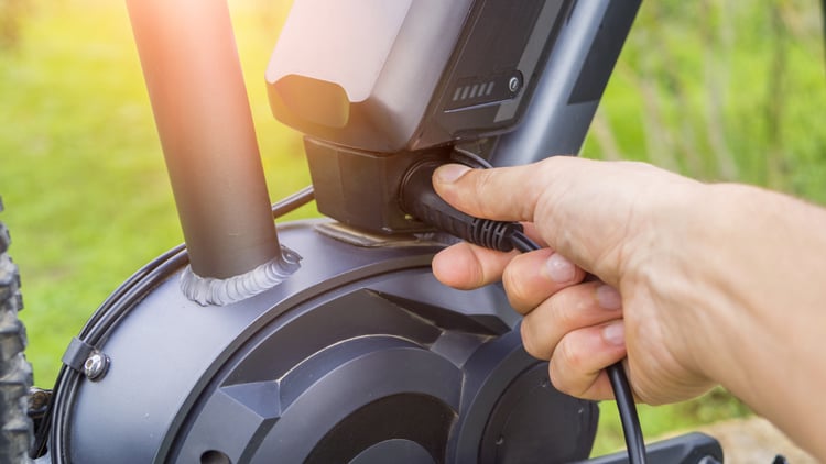 E-Bike richtig laden Nahaufnahme einer Hand, die ein Ladekabel in den Akku eines E-Bikes einsteckt; Fokus auf dem Anschlussbereich am Fahrradrahmen, im Hintergrund unscharfe grüne Außenumgebung.