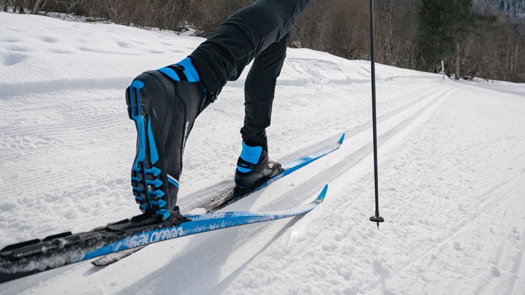 Nahaufnahme eines Langläufers auf einem verschneiten Weg. Der Fokus liegt auf den schwarzen Skischuhen mit blauen Akzenten und den dazu passenden Skiern, von denen einer angehoben ist. Ein Skistock ist im Hintergrund sichtbar