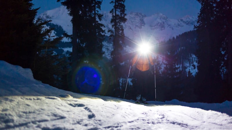 Eine Person geht bei einer nächtlichen Skitour mit eingeschalteter Stirnlampe durch den verschneiten Wald.