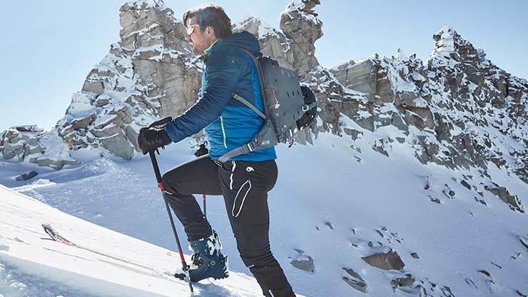 Get Green Person steigt mit Skistöcken einen verschneiten Berg hinauf, gekleidet in blaue Winterjacke, schwarze Hose und mit Rucksack. Im Hintergrund sind schroffe, schneebedeckte Berggipfel unter klarem blauem Himmel zu sehen