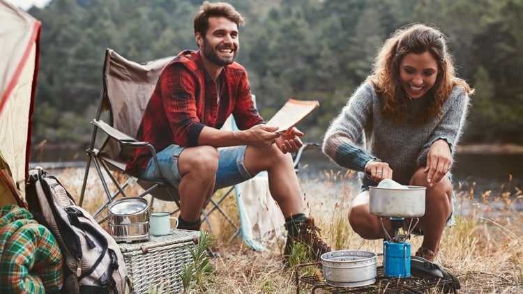 Zwei Personen beim Camping im Freien: Eine sitzt in einem Klappstuhl mit rot-karierter Bluse und Jeansshorts und hält ein Blatt Papier, die andere hockt am Campingkocher in grauem Pullover. Um sie herum stehen Zelt, Metallbecher und weitere Campingutensilien. Im Hintergrund Bäume und ein Gewässer.