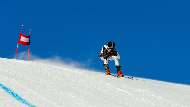 Skifahrer in Rennanzug und Helm fährt in geduckter Haltung mit hoher Geschwindigkeit eine verschneite Piste hinab. Links im Bild ist eine rot-weiße Flagge als Teil der Rennstrecke zu sehen. Der Himmel ist klar und blau