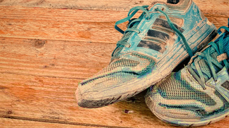 Ein Paar abgenutzte, blaue Sportschuhe mit schwarzen Streifen auf einer Holzoberfläche. Die Schuhe zeigen deutliche Gebrauchsspuren und sind sichtbar verschmutzt