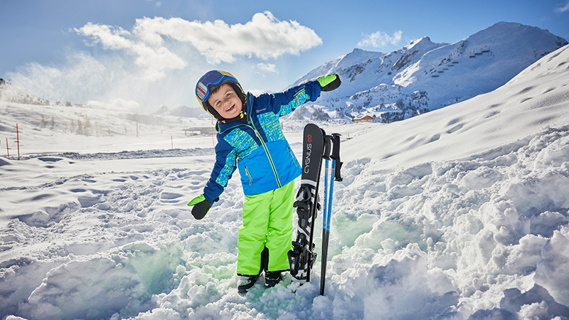 Ein Kind in bunter Skibekleidung steht lachend im Schnee und lehnt sich an seine Ski.
