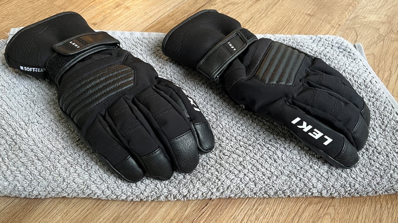 Ein Paar schwarze LEKI-Skihandschuhe liegt nebeneinander auf einem grauen Handtuch, ausgebreitet auf einem Holzboden zum Trocknen nach dem Einsatz.
