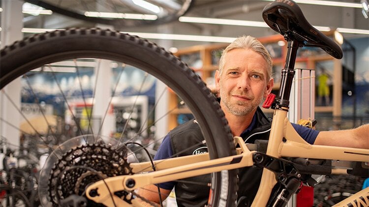 Bike Service Termin Bike Service Termin. Ein Mann steht hinter einem Fahrrad mit braunem Rahmen. Er schut direkt in die Kamera. Im Hintergrund sind weitere Fahrräder.