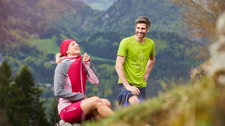 Zwei Personen in bergiger Umgebung im Freien. Eine sitzt auf einem Felsen und trägt ein rot-graues Outfit mit roter Kopfbedeckung. Die andere steht daneben in grünem Shirt und blauer Shorts. Im Hintergrund sind grüne Bäume und Berge zu sehen
