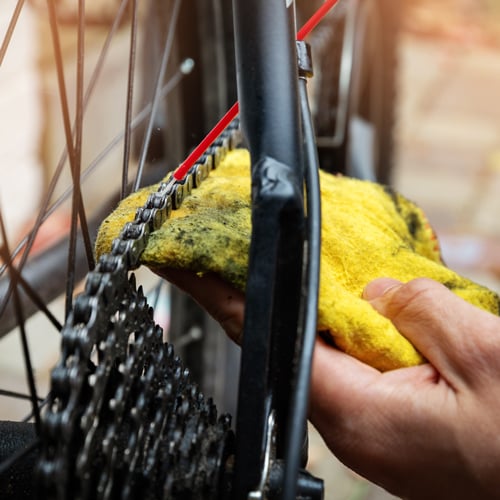 Nahaufnahme einer Person, die mit einem gelben Tuch die Fahrradkette reinigt und gleichzeitig mit einem Spray samt rotem Röhrchen Schmiermittel aufträgt.