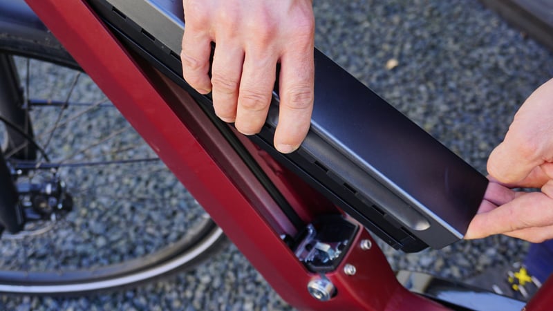 Nahaufnahme von Händen, die den Akku eines roten E-Bikes entfernen. Der Akku ist schwarz, im Hintergrund ist ein Boden aus Kies oder kleinen Steinen zu sehen.