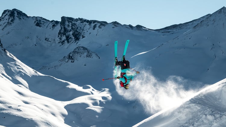 Skifahrer führt einen Rückwertssalto in einem verschneiten Gebirgsgebiet aus. Er ist kopfüber in der Luft mit nach oben gerichteten Skiern, während Schnee aufgewirbelt wird. Im Hintergrund sind schneebedeckte Berge unter klarem, blauem Himmel zu sehen