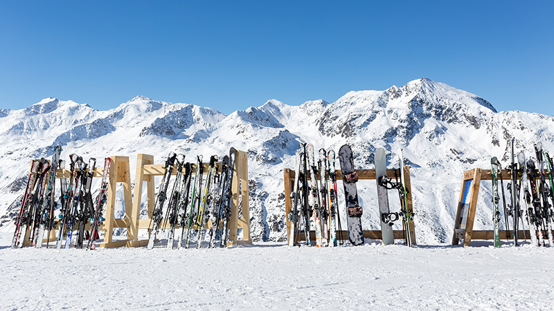 Mehrere Paar Ski sind nebeneinander in einer Halterung aufgestellt, im Hintergrund sind verschneite Berge zu sehen.