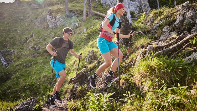 Zwei Personen wandern auf einem felsigen Bergpfad. Sie tragen Rucksäcke, Shorts und Sportschuhe und benutzen Trekkingstöcke. Im Hintergrund sind Grasflächen, Bäume und bergiges Gelände zu sehen