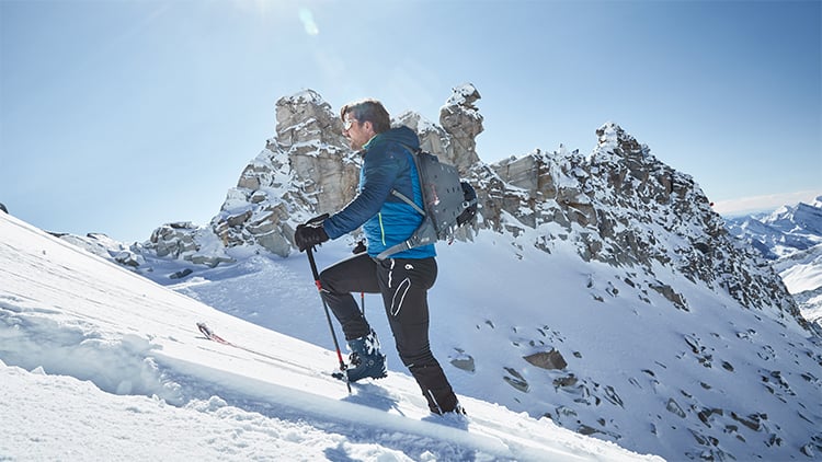 Person steigt mit Skistöcken einen verschneiten Berg hinauf, gekleidet in blaue Winterjacke, schwarze Hose und mit Rucksack. Im Hintergrund sind schroffe, schneebedeckte Berggipfel unter klarem blauem Himmel zu sehen
