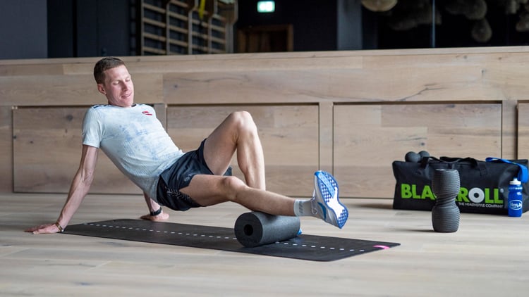 Laufberater Person in Sportkleidung nutzt eine Faszienrolle zur Selbstmassage des hinteren Oberschenkels auf einer Trainingsmatte; daneben liegen eine BLACKROLL-Tasche, eine weitere Rolle und eine Wasserflasche