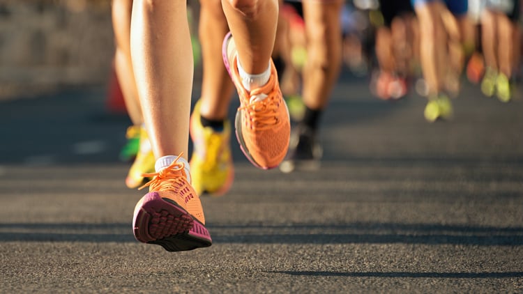 Gruppe von Läuferinnen und Läufern bei einem Rennen auf einer asphaltierten Straße. Der Fokus liegt auf den Beinen und bunten Laufschuhen in Farben wie Orange und Gelb. Der Hintergrund ist leicht verschwommen und vermittelt Bewegung