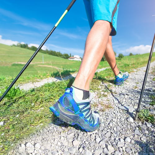 Person wandert mit Trekkingstöcken auf einem Schotterweg, Fokus auf blaue Wanderschuhe vor grünem Gras und blauem Himmel.