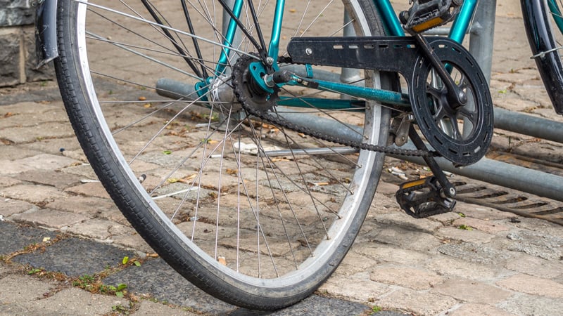 Nahaufnahme des hinteren Laufrads mit einem Achter eines türkisfarbenen Fahrrads. Das Fahrrad steht auf einem gepflasterten Untergrund, im Hintergrund sind Fahrradständer zu sehen. Kette, Pedale und Teile des Schaltwerks sind ebenfalls sichtbar.